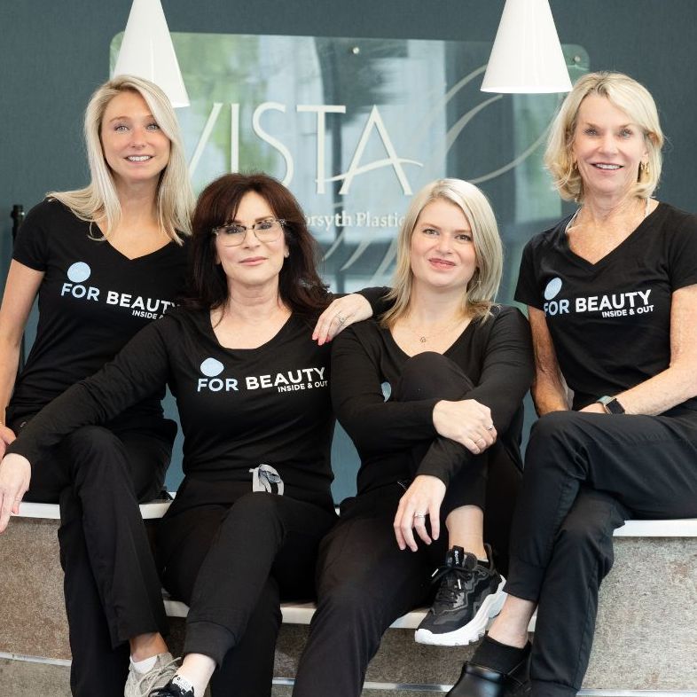 The VISTA Med Spa team sit together on the front desk ledge inside the practice, smiling and wearing matching black "FOR BEAUTY" shirts. The modern reception area with the VISTA logo is visible in the background.